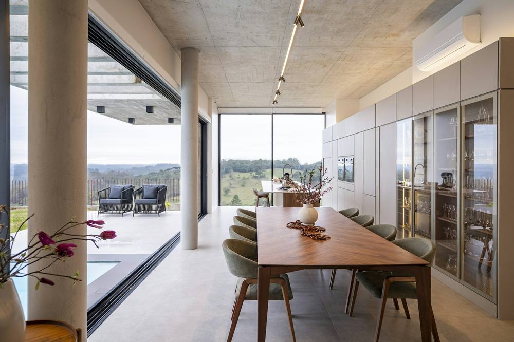 A long kitchen and dining room with a pale stone floor opening through full-height glazing to a terrace and hills
