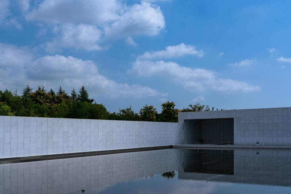 A still reflecting pool edged by a long pale stone wall under a blue sky with clouds
