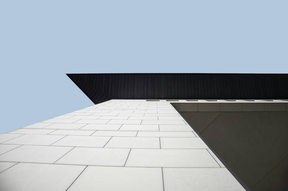 Large pale stone panels on a building corner below a dark overhang against a blue sky