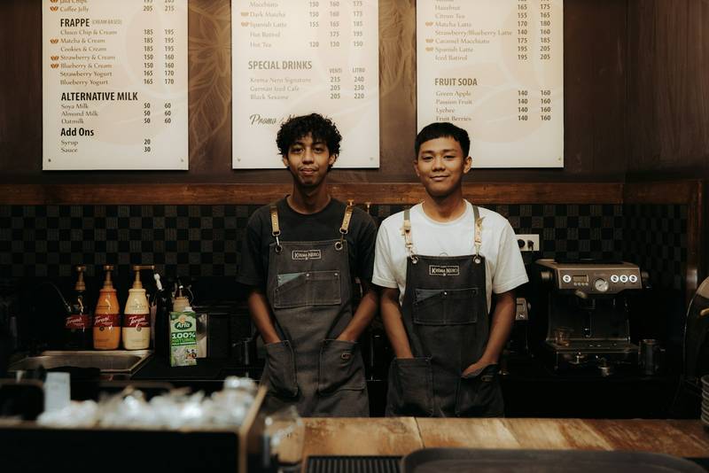 Two baristas in aprons smile behind a cafe counter