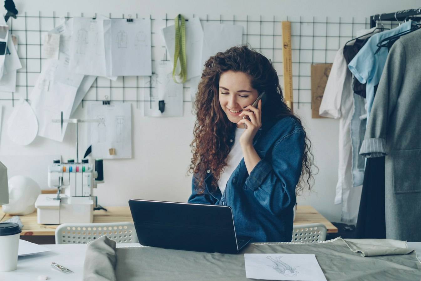A clothing designer smiles on the phone at her studio workbench, laptop open beside her sketches
