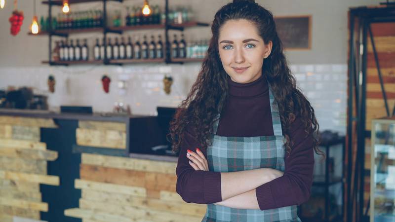 A coffee shop owner in an apron stands with arms folded in front of her counter